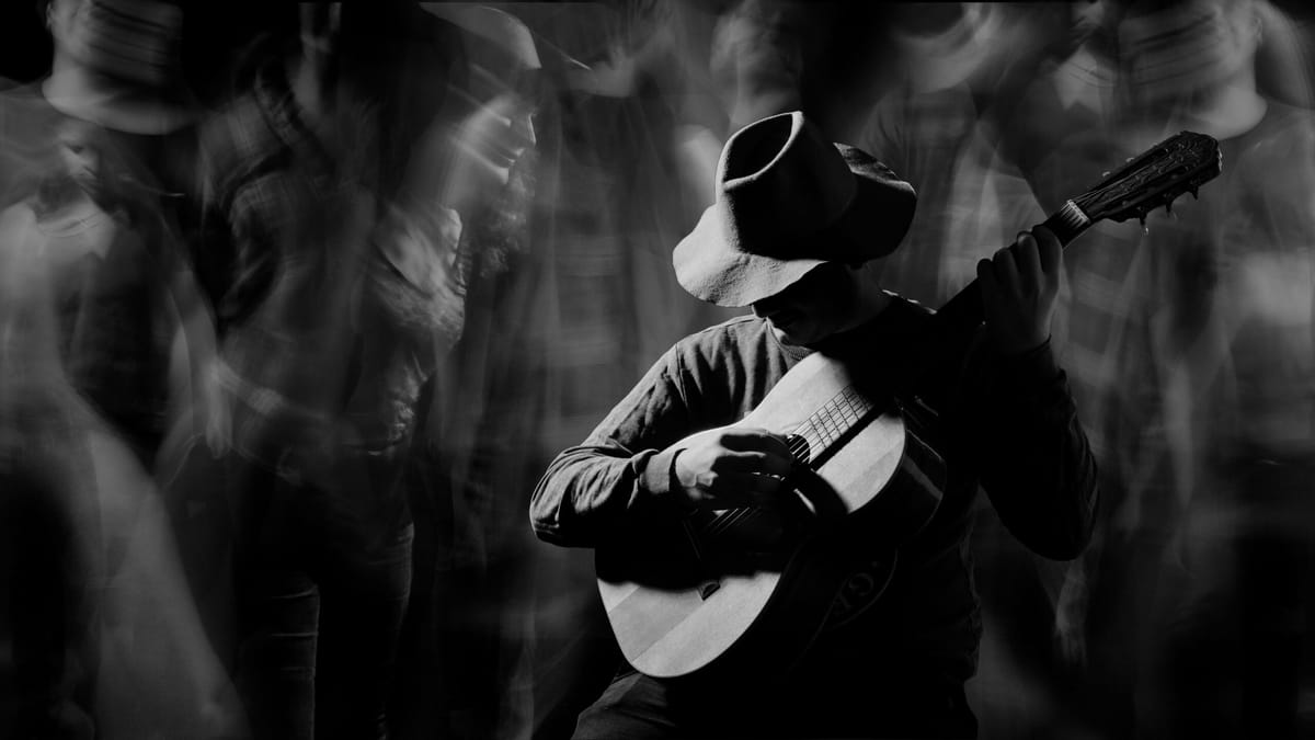 Foto em preto e branco de um músico segurando um violão cercado por borrões de pessoas. Seu rosto está coberto por um chapéu,